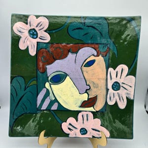 May include: Square ceramic plate with a painted abstract face in shades of purple, peach, and yellow. The plate is decorated with pink flowers and green leaves on a dark green background. The plate is displayed on a wooden stand.
