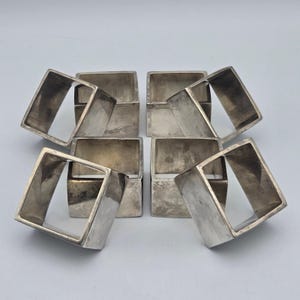 May include: A set of four square napkin rings made of polished silver-toned metal. Each ring features a hollow center and a smooth, reflective surface with subtle variations in tone, arranged in a symmetrical two-by-two grid formation.