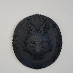 May include: A matte black wall plaque featuring a sculpted fox head in the centre. The fox is surrounded by an ornate, circular frame with decorative scrollwork details along the outer edge. The entire piece is monochromatic and solid black.
