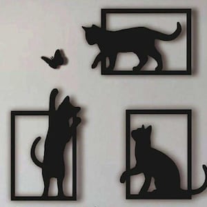 May include: A set of three black wall-mounted silhouettes featuring two cats inside rectangular frames, one cat reaching upward towards a butterfly, and one cat sitting inside a frame. The designs are flat, solid black shapes with clean edges.