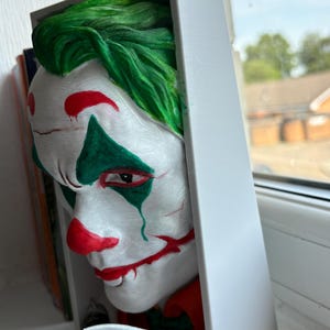 以下が含まれることがあります： ジョーカーの顔の3Dプリント彫刻。緑色の髪、鼻と口の赤いアクセント、白い顔が特徴です。彫刻は白い長方形のフレームに部分的に囲まれており、銀色のナイフが見えます。