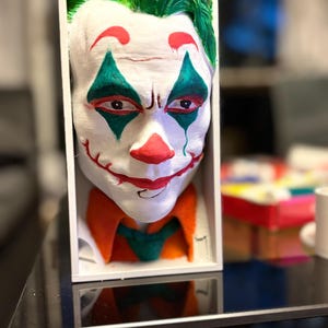 以下が含まれることがあります： 白い手描きのジョーカーのマスクで、白い箱に額装されています。マスクは緑色の髪、目と口の周りの赤いアクセント、緑とオレンジの襟が特徴です。マスクは象徴的な悪役の詳細な表現です。