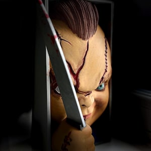 Peut inclure: Une sculpture murale en trois dimensions représentant une tête de poupée avec une crête brune texturée, des cicatrices faciales visibles et des yeux bleus. Une main sort du cadre tenant un couteau argenté avec des taches de sang rouge.