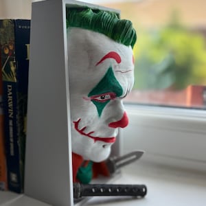 以下が含まれることがあります： ジョーカーをテーマにしたデザインの白い本棚。本棚には、緑の髪、赤い唇、緑と赤のペイントが施されたジョーカーの顔が彫刻されています。小さなナイフと黒い物体も表示されています。