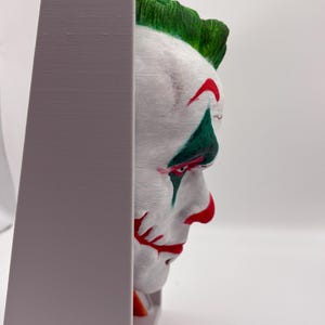 以下が含まれることがあります： ジョーカーの顔の3Dプリント彫刻。白い肌、赤い唇、緑の髪が特徴です。彫刻は灰色のフレームに収められています。ジョーカーの顔は鮮やかな色で塗装されています。ユニークな芸術作品です。