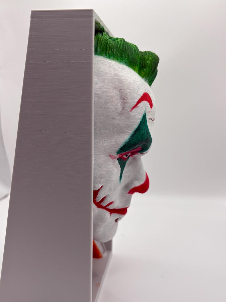 以下が含まれることがあります： ジョーカーの顔の3Dプリント彫刻。白い肌、赤い唇、緑の髪が特徴です。彫刻は灰色のフレームに収められています。ジョーカーの顔は鮮やかな色で塗装されています。ユニークな芸術作品です。