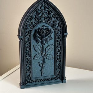 Puede incluir: Una placa decorativa negra en forma de arco que presenta un relieve central de una rosa con hojas y tallo. La rosa está enmarcada por un intrincado patrón de celosía calada que recuerda a la tracería gótica, todo en acabado negro mate.