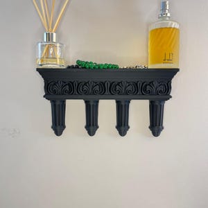 以下が含まれることがあります： 彫刻が施されたデザインの黒い装飾的な壁棚。ガラス製のディフューザー、香水瓶、緑色のビーズが置かれています。棚は4本の支柱で支えられており、明るい色の壁に取り付けられています。
