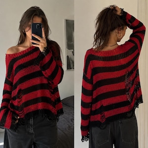 Könnte beinhalten: Ein langärmeliger, locker sitzender Pullover mit horizontalen roten und schwarzen Streifen. Das Strickkleidungsstück hat einen weiten, offenen Ausschnitt und weist abgenutzte, ausgefranste Details am gesamten Stoff auf, einschließlich am Saum und an den Ärmelbündchen.
