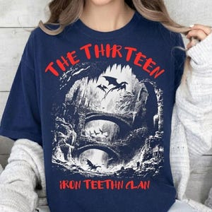 May include: Navy blue t-shirt with a graphic featuring a fantasy scene with bridges, dragons, and the text "THE THIRTEEN IRON TEEHTN CLAN" in red. The design is a detailed illustration with a gothic aesthetic.