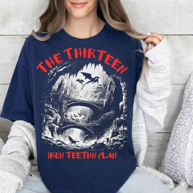 May include: Navy blue t-shirt with a graphic featuring a fantasy scene with bridges, dragons, and the text "THE THIRTEEN IRON TEEHTN CLAN" in red. The design is a detailed illustration with a gothic aesthetic.