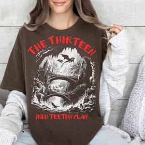 May include: Brown t-shirt with a graphic featuring a fantasy scene with dragons, bridges, and the text "THE THIRTEEN IRON TEEHTN CLAN" in red. The design is a detailed black and white illustration.