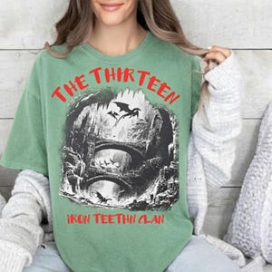 May include: Sage green t-shirt with a graphic featuring a fantasy scene with dragons, bridges, and the text "THE THIRTEEN IRON TEEHTN CLAN" in red. The shirt has a vintage, worn look.