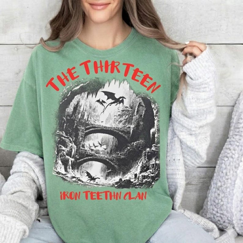 May include: Sage green t-shirt with a graphic featuring a fantasy scene with dragons, bridges, and the text "THE THIRTEEN IRON TEEHTN CLAN" in red. The shirt has a vintage, worn look.