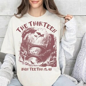 May include: Cream-colored t-shirt featuring a maroon graphic of a fantasy landscape with bridges, dragons, and the text "THE THIRTEEN" and "IRON TEETHN CLAN". The design is a vintage-style illustration.