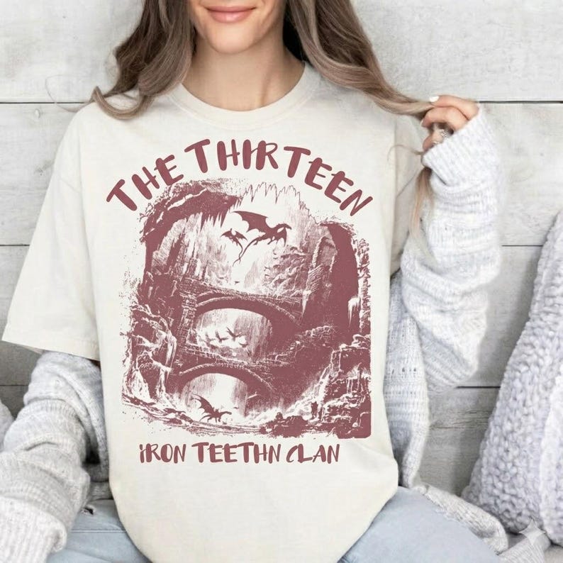 May include: Cream-colored t-shirt featuring a maroon graphic of a fantasy landscape with bridges, dragons, and the text "THE THIRTEEN" and "IRON TEETHN CLAN". The design is a vintage-style illustration.