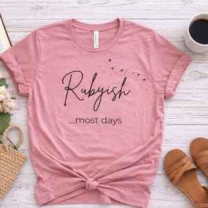 Puede incluir: Tres camisetas en diferentes colores: rosa empolvado, verde salvia y gris claro. Cada camiseta tiene la palabra "Rubbish" en cursiva con la frase "...most days" debajo. Las camisetas están diseñadas con sandalias, café y libros.