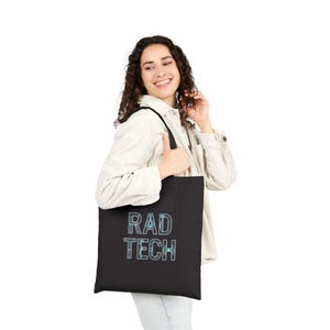 Puede incluir: Bolso tote negro con las palabras "RAD TECH" en azul claro. La persona lleva una camisa blanca y vaqueros azules.