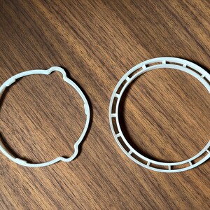 May include: Two white, circular objects are displayed on a wooden surface. One is a solid circle with a scalloped edge. The other is a ring with rectangular cutouts. Both objects appear to be made of plastic.