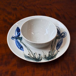 May include: A white ceramic bowl centered on a matching white ceramic plate. The plate features hand-painted blue and grey floral motifs with thin, dark stems and delicate petals arranged around the perimeter of the bowl's base.