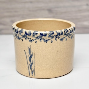 May include: A beige stoneware crock with a blue floral design around the rim and a blue wheat stalk illustration on the side. The cylindrical container is a classic piece of kitchenware.