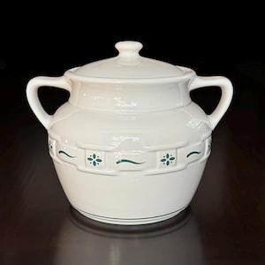May include: A cream-colored ceramic sugar bowl with a matching lid and two side handles. The body features a raised basket-weave texture around the center, accented with small green floral motifs and thin horizontal green wavy lines.