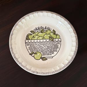May include: A round ceramic pie dish with a fluted edge and brown line detailing. The center features a green apple illustration and the text "apple pie" above a printed recipe with instructions for baking a two-crust apple pie in the oven.