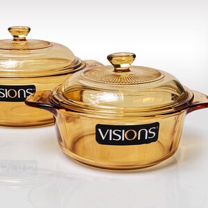 May include: Two amber-colored glass cooking pots with matching lids. Each pot features integrated side handles and a black rectangular label on the front with the white text "VISIONS" and an orange circle accent. The lids have a radial pattern.