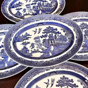 May include: A collection of oval ceramic plates featuring a classic blue and white willow pattern. Each plate displays a detailed transfer print of a landscape with pagodas, willow trees, a bridge, and birds in flight, framed by a decorative border.