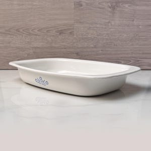 May include: White rectangular baking dish with a blue floral design on the side. The dish has a slightly raised rim and is made of ceramic. The background is a light wood grain.