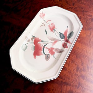 May include: An octagonal cream-colored serving platter featuring a floral pattern with muted red and pink blossoms, green leaves, and thin stems. The platter has a smooth, glossy finish and a thin, dark-colored rim accenting its geometric shape.