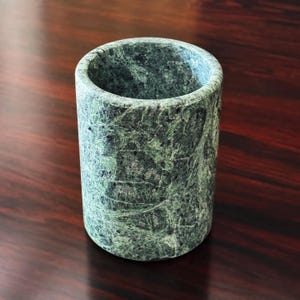 May include: A cylindrical vessel crafted from polished green stone featuring intricate dark veining and a smooth, mottled texture throughout its exterior surface. The container has a wide, open top and a solid, uniform structure with a deep, hollow interior.