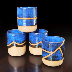 May include: A set of four cylindrical ceramic cups featuring a deep blue glaze on the upper portion and a natural beige clay base. Each cup displays distinct tan-colored line patterns, including horizontal bands, chevron shapes, and geometric segments.