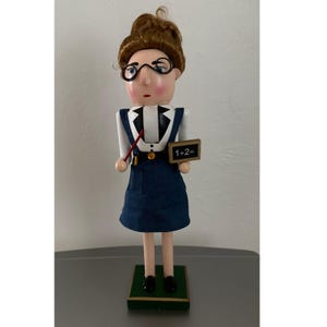 May include: A wooden nutcracker in the shape of a teacher, wearing a white shirt, blue jumper, and black shoes. The nutcracker has brown hair, glasses, and holds a pointer and a chalkboard that reads "1+2=". The base is green.