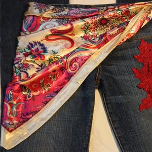 May include: Dark blue denim jeans featuring a vibrant, multi-colored satin scarf draped across the waist. The scarf displays a detailed paisley and floral pattern in shades of pink, red, gold, and blue. A red lace floral applique is attached to the side.
