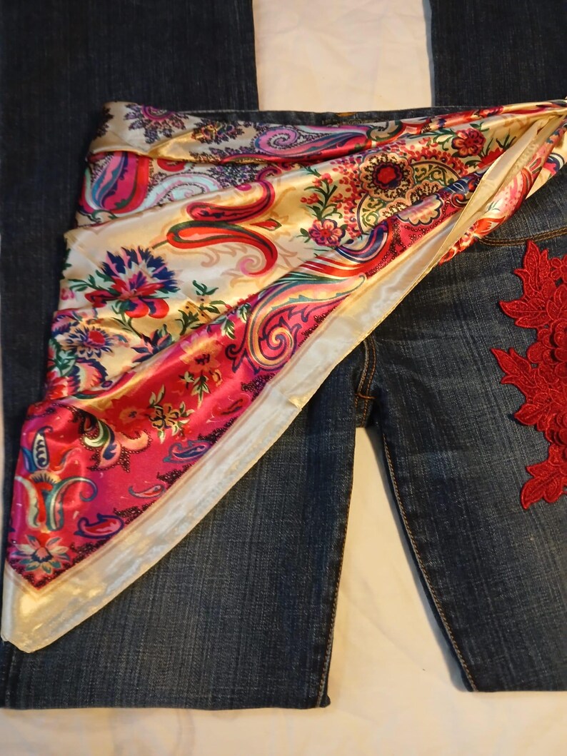 May include: Dark blue denim jeans featuring a vibrant, multi-colored satin scarf draped across the waist. The scarf displays a detailed paisley and floral pattern in shades of pink, red, gold, and blue. A red lace floral applique is attached to the side.