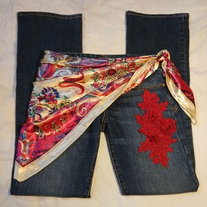 May include: Dark blue denim jeans featuring a vibrant, multi-colored paisley print silk scarf tied around the waist. The jeans are adorned with a prominent, intricate red floral lace appliqu&eacute; stitched onto the upper front of the right leg.