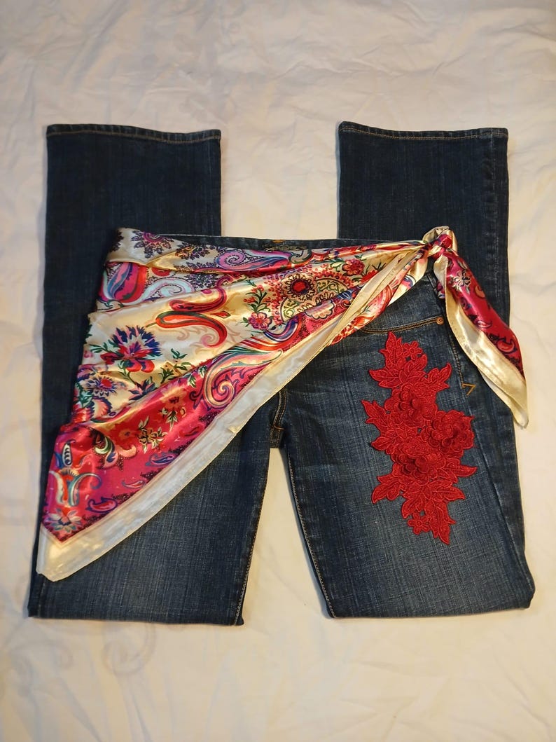 May include: Dark blue denim jeans featuring a vibrant, multi-colored paisley print silk scarf tied around the waist. The jeans are adorned with a prominent, intricate red floral lace appliqu&eacute; stitched onto the upper front of the right leg.