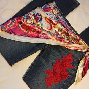 May include: Dark blue denim jeans featuring a large, intricate red floral lace appliqu&eacute; on the upper thigh area. A vibrant, multi-colored satin scarf with a paisley and floral pattern in shades of pink, red, and cream is draped across the jeans.