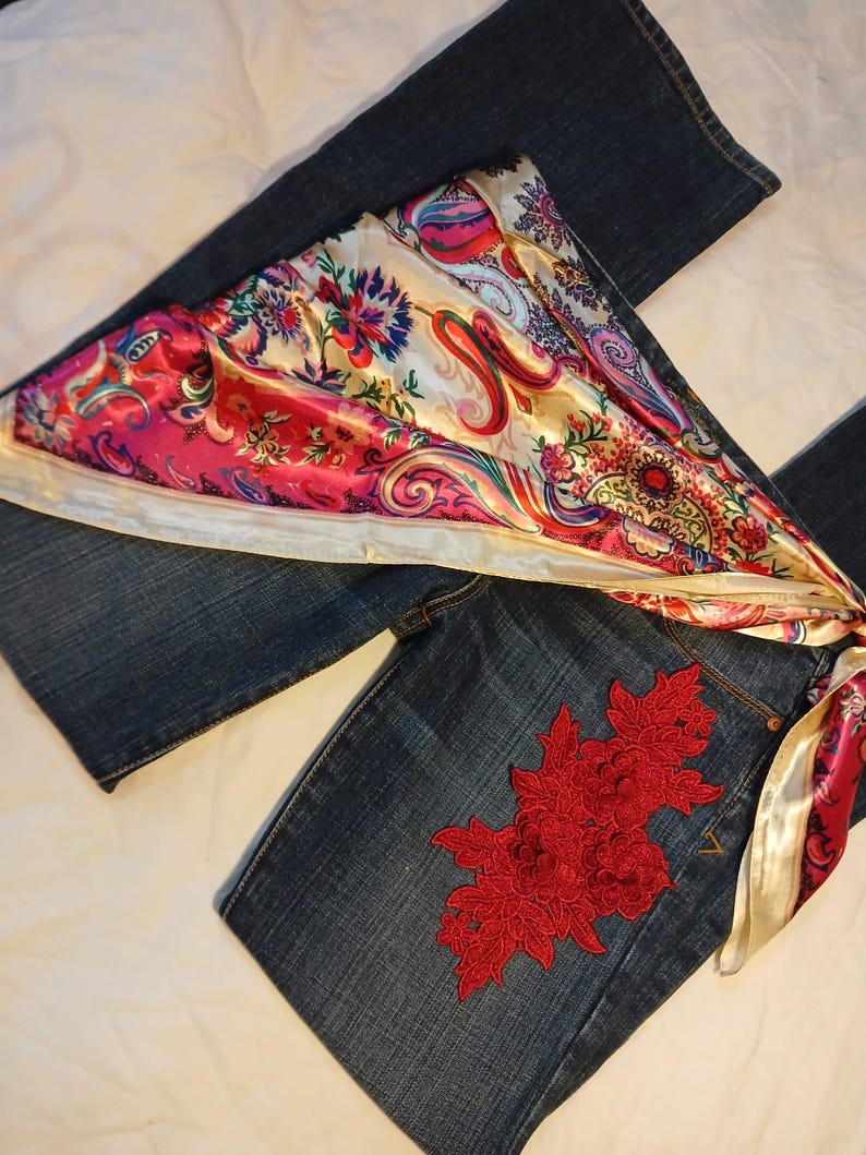 May include: Dark blue denim jeans featuring a large, intricate red floral lace appliqu&eacute; on the upper thigh area. A vibrant, multi-colored satin scarf with a paisley and floral pattern in shades of pink, red, and cream is draped across the jeans.
