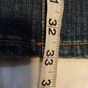 May include: A pair of dark blue denim jeans featuring thick, contrasting orange stitching along the seams. A white measuring tape is positioned vertically against the fabric, showing measurements in inches ranging from 31 to 34 inches.