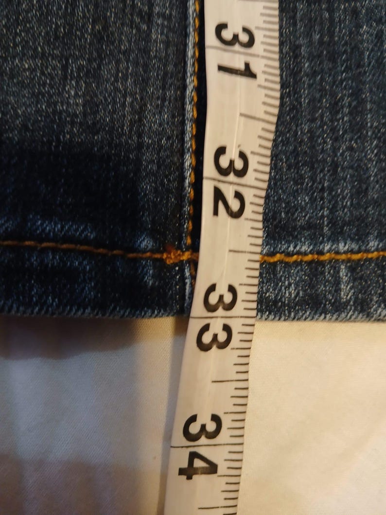 May include: A pair of dark blue denim jeans featuring thick, contrasting orange stitching along the seams. A white measuring tape is positioned vertically against the fabric, showing measurements in inches ranging from 31 to 34 inches.