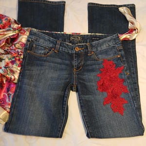 May include: Dark blue denim boot cut jeans featuring a prominent red floral lace appliqu&eacute; on the left thigh. The waistband includes a colorful paisley patterned fabric trim and a Seven7 brand label. The jeans have a classic button and zipper fly.