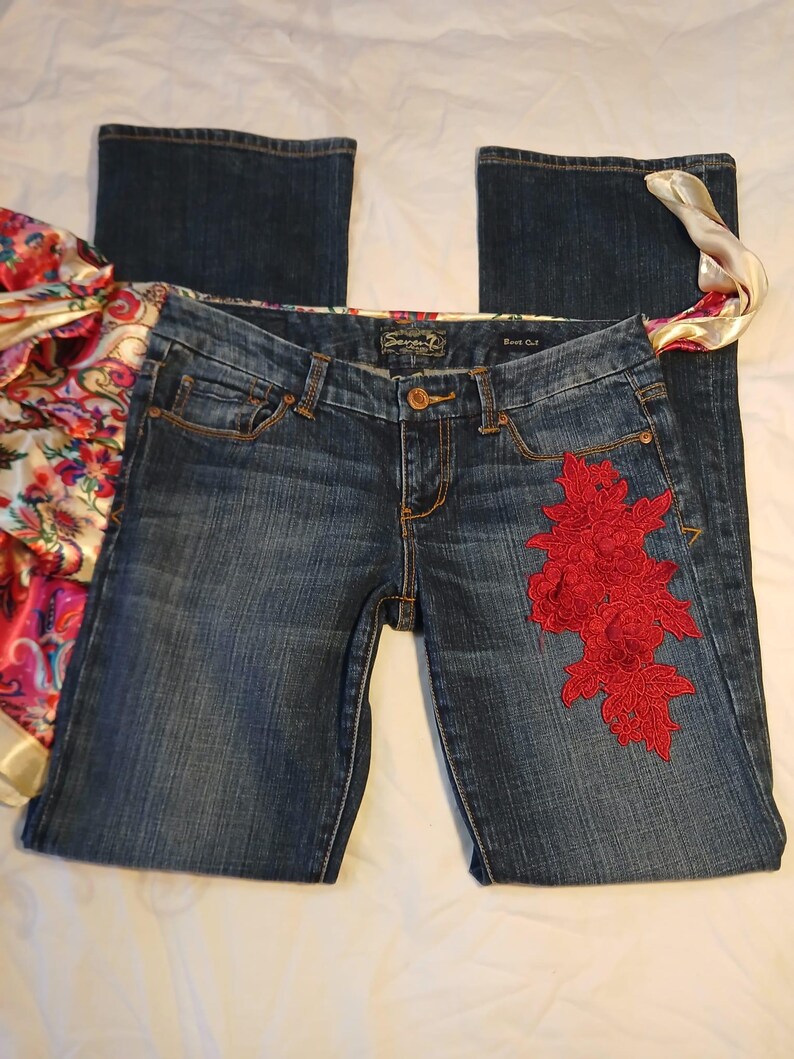 May include: Dark blue denim boot cut jeans featuring a prominent red floral lace appliqu&eacute; on the left thigh. The waistband includes a colorful paisley patterned fabric trim and a Seven7 brand label. The jeans have a classic button and zipper fly.