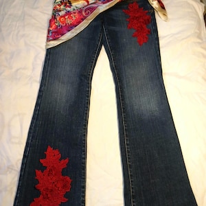 May include: Dark blue denim jeans featuring a vibrant, multicolored paisley print satin sash tied at the waist. The jeans are embellished with large, intricate red floral lace appliqu&eacute;s positioned on the upper left thigh and the lower right leg.