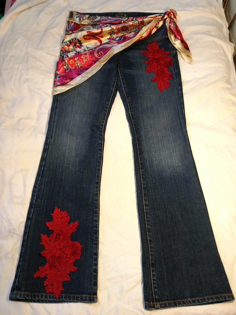 May include: Dark blue denim jeans featuring a vibrant, multicolored paisley print satin sash tied at the waist. The jeans are embellished with large, intricate red floral lace appliqu&eacute;s positioned on the upper left thigh and the lower right leg.
