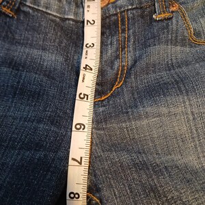 May include: Blue denim jeans featuring a copper-colored button and orange contrast stitching. A white measuring tape is placed vertically over the front, showing a length measurement of approximately 9 inches from the waistband to the crotch seam.