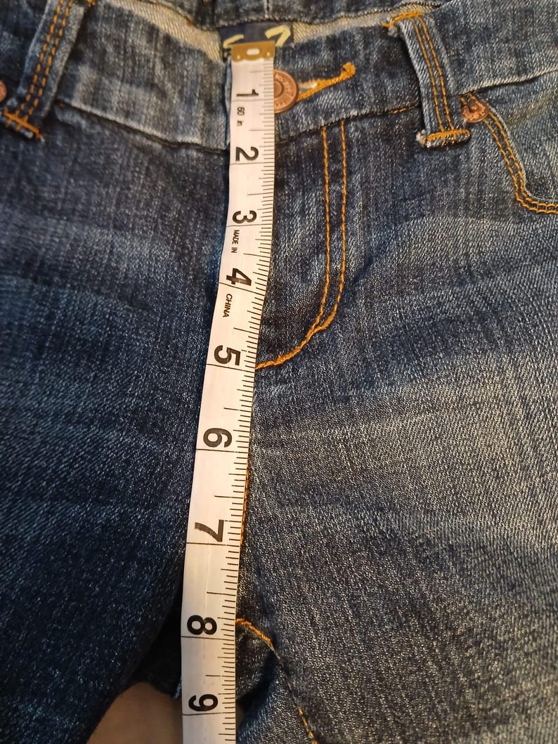 May include: Blue denim jeans featuring a copper-colored button and orange contrast stitching. A white measuring tape is placed vertically over the front, showing a length measurement of approximately 9 inches from the waistband to the crotch seam.
