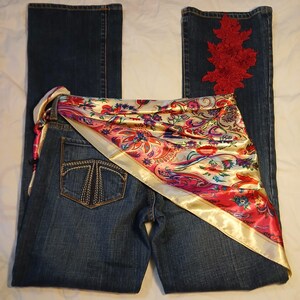 May include: Dark blue denim jeans featuring a back pocket with decorative stitching and a red floral lace appliqu&eacute; on the right leg. A satin scarf with a colorful paisley and floral pattern in shades of pink, red, and gold is draped across the waist.