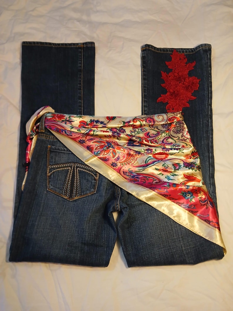 May include: Dark blue denim jeans featuring a back pocket with decorative stitching and a red floral lace appliqu&eacute; on the right leg. A satin scarf with a colorful paisley and floral pattern in shades of pink, red, and gold is draped across the waist.